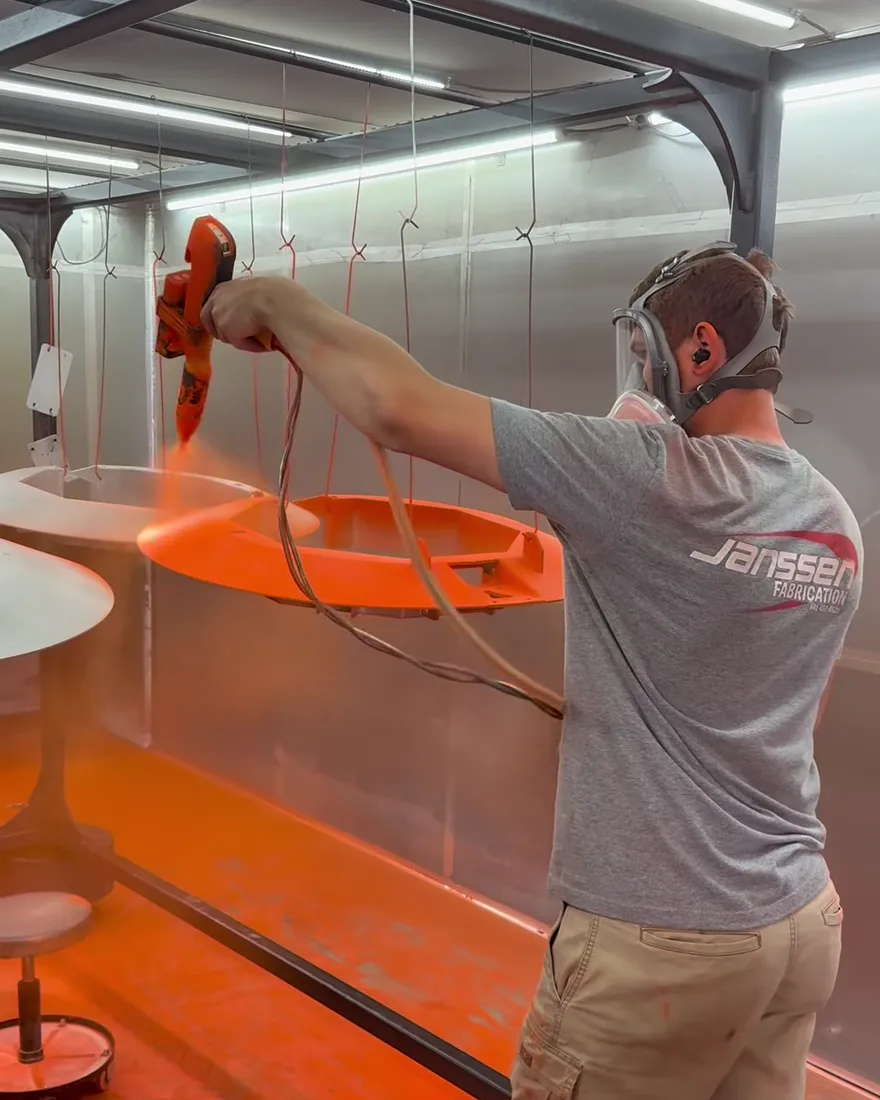 A Janssen Fabrication team member in a company shirt sprays orange powder onto a hanging frame in the booth.