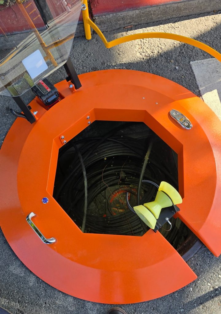 Top-down view of Light Ring manhole cover ring with aluminum frame, integrated hose roller, and drill battery deployed over open manhole.